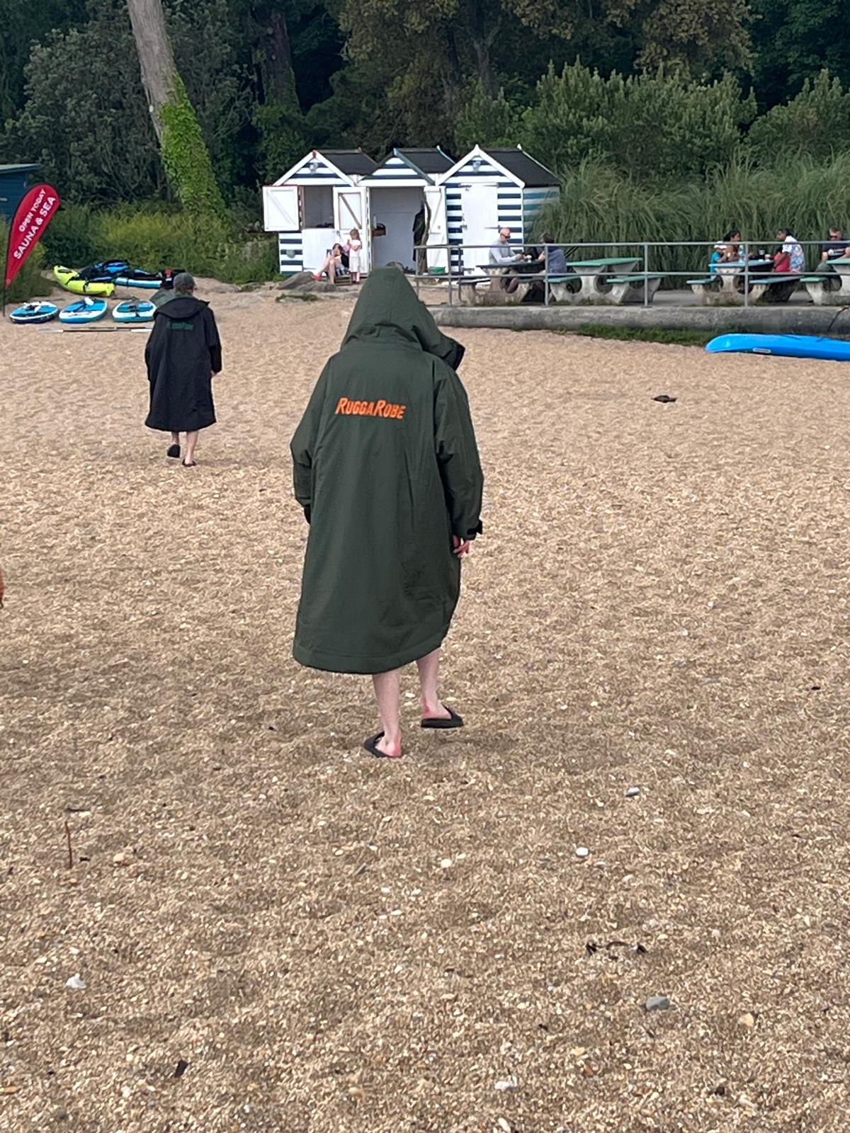 Person wearing a green ruggarobe with text on the back, standing on a sandy area with a small building and people in the background.