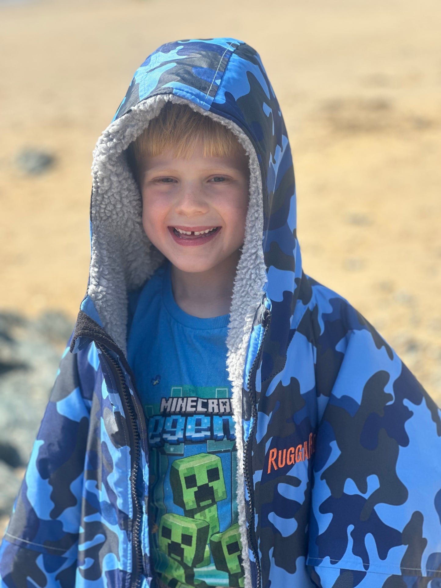 Child wearing a blue camouflage ruggarobe with a Minecraft shirt underneath, standing on a sandy background.