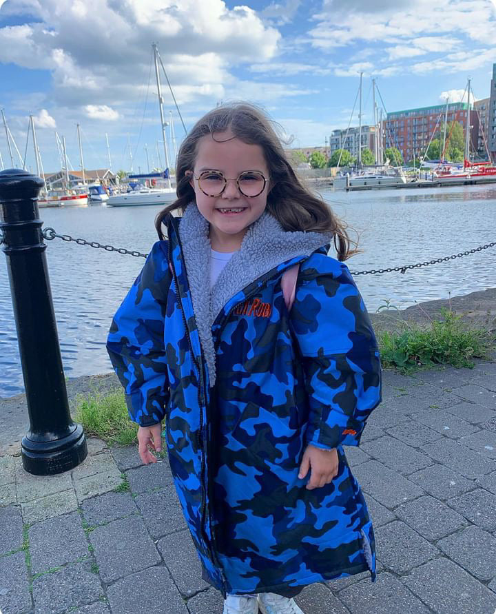 Child wearing a blue camouflage ruggarobe by a waterfront with boats and buildings in the background.