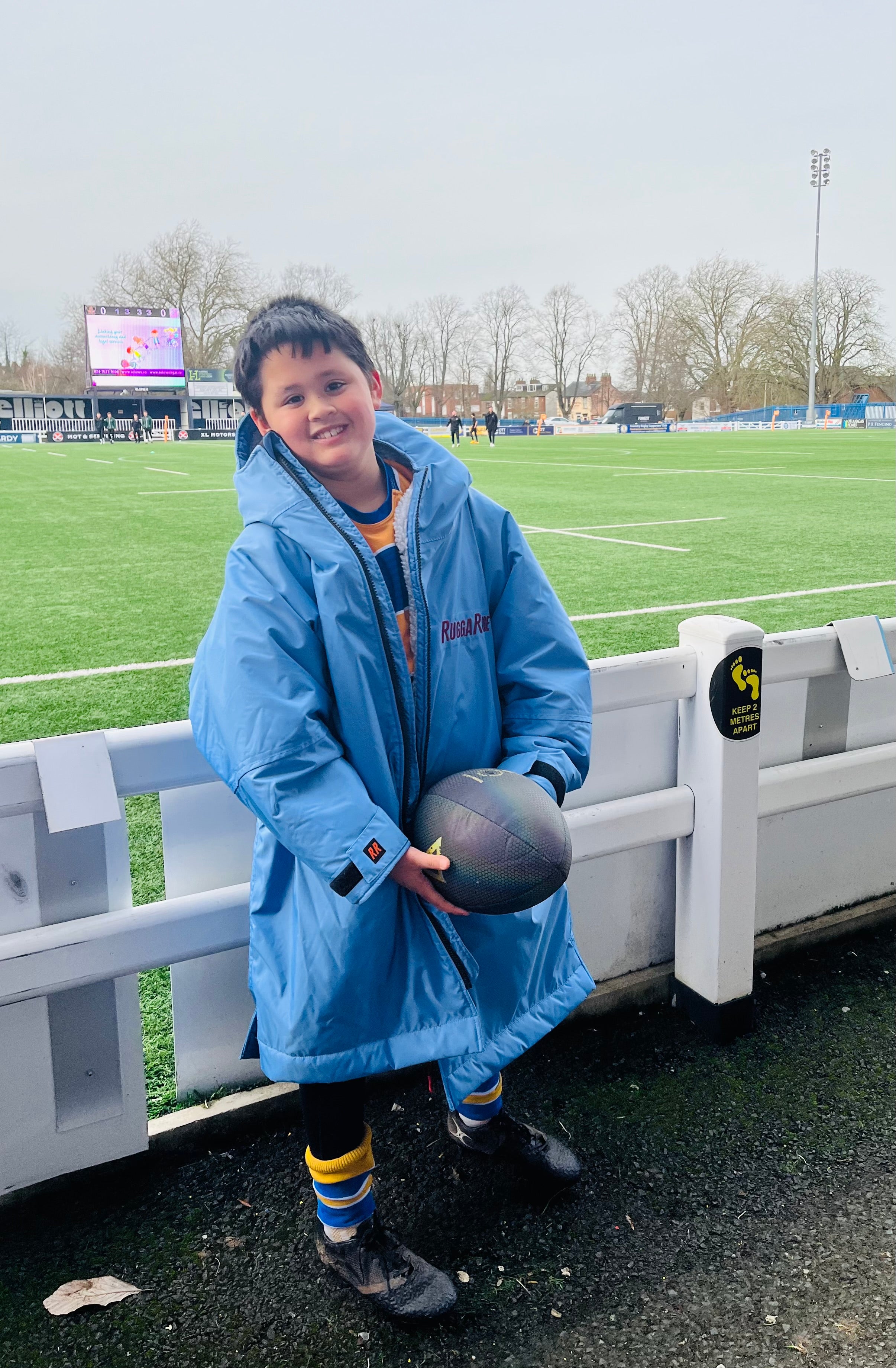 Child in a blue coat holding a rugby ball on a sports field