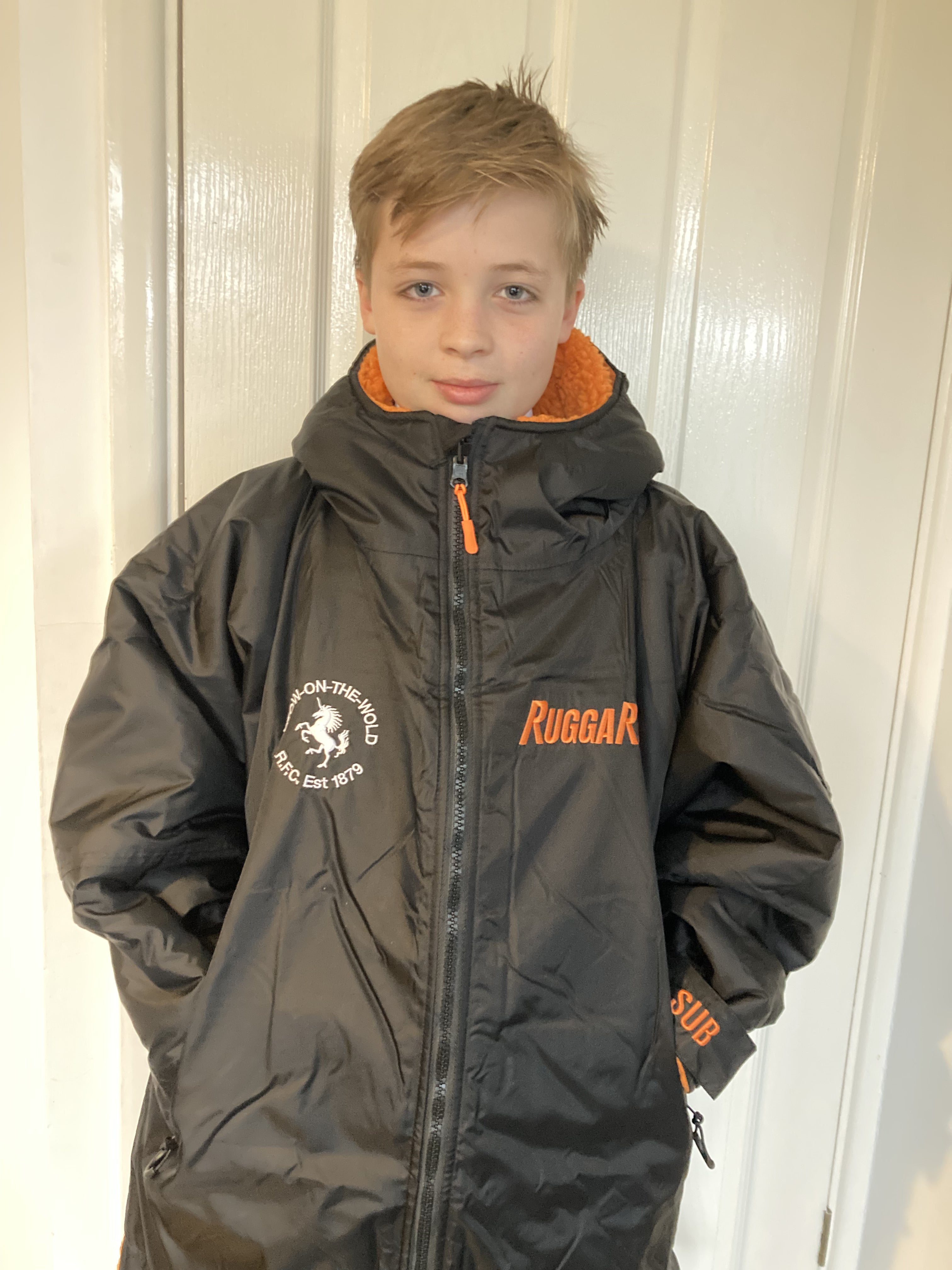 A person wearing a black rugge robe change robe with orange fleece lining, featuring a hood and a front zip, standing indoors.