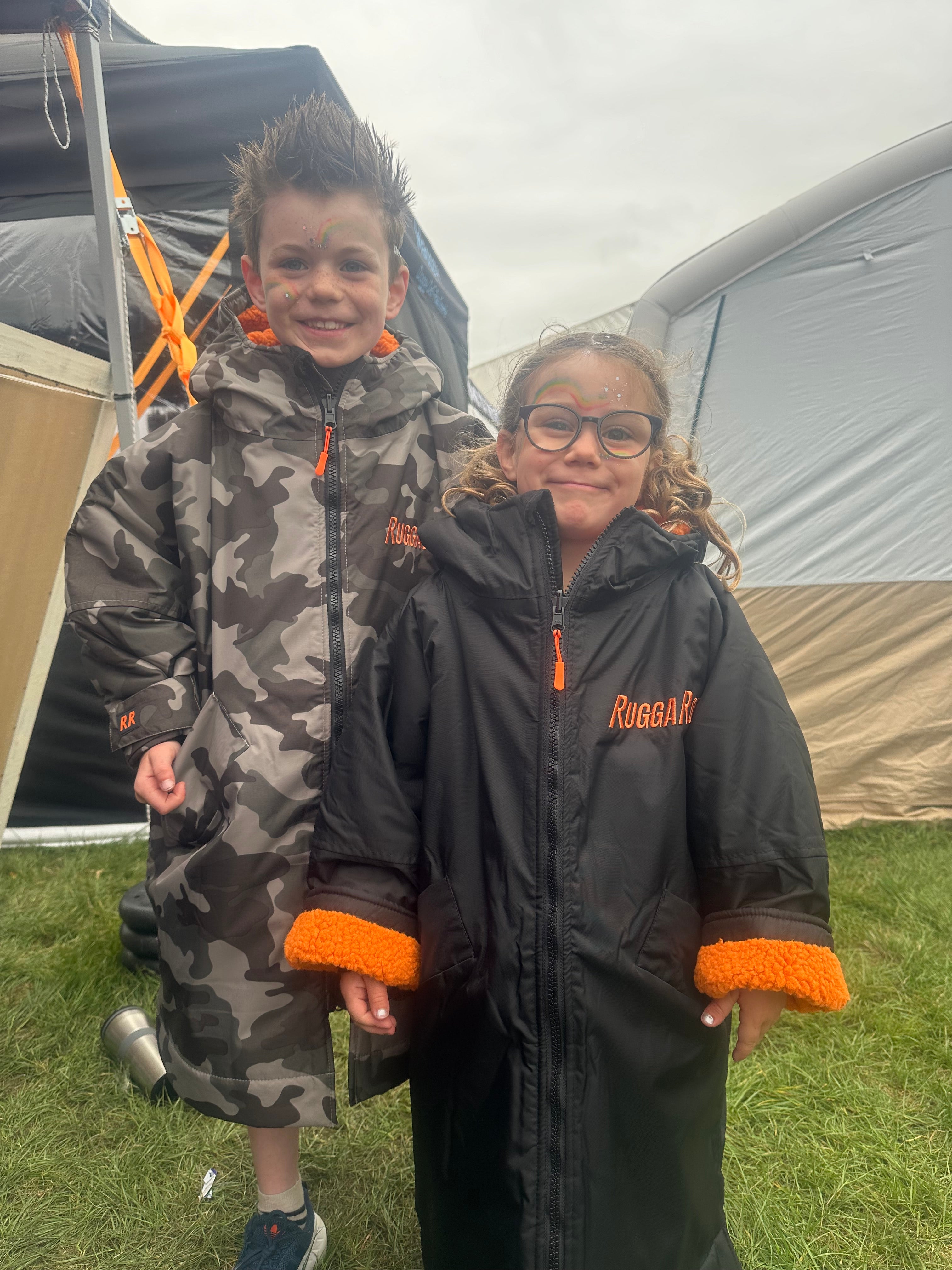 Two children wearing black and orange RuggaRobe change robes standing outdoors near a tent.