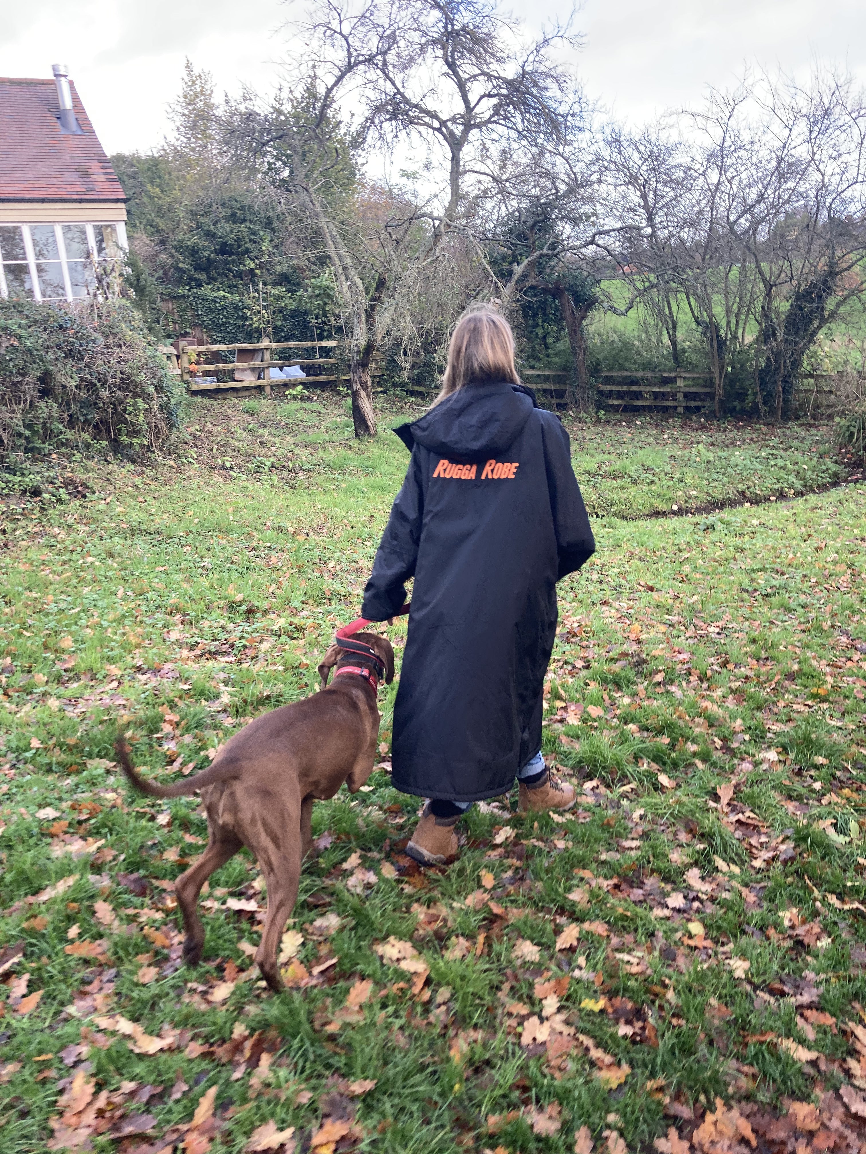 Dog walker wearing a black changerobe with orange lining and 'RuggaRobe' branding. Walking with dog in field