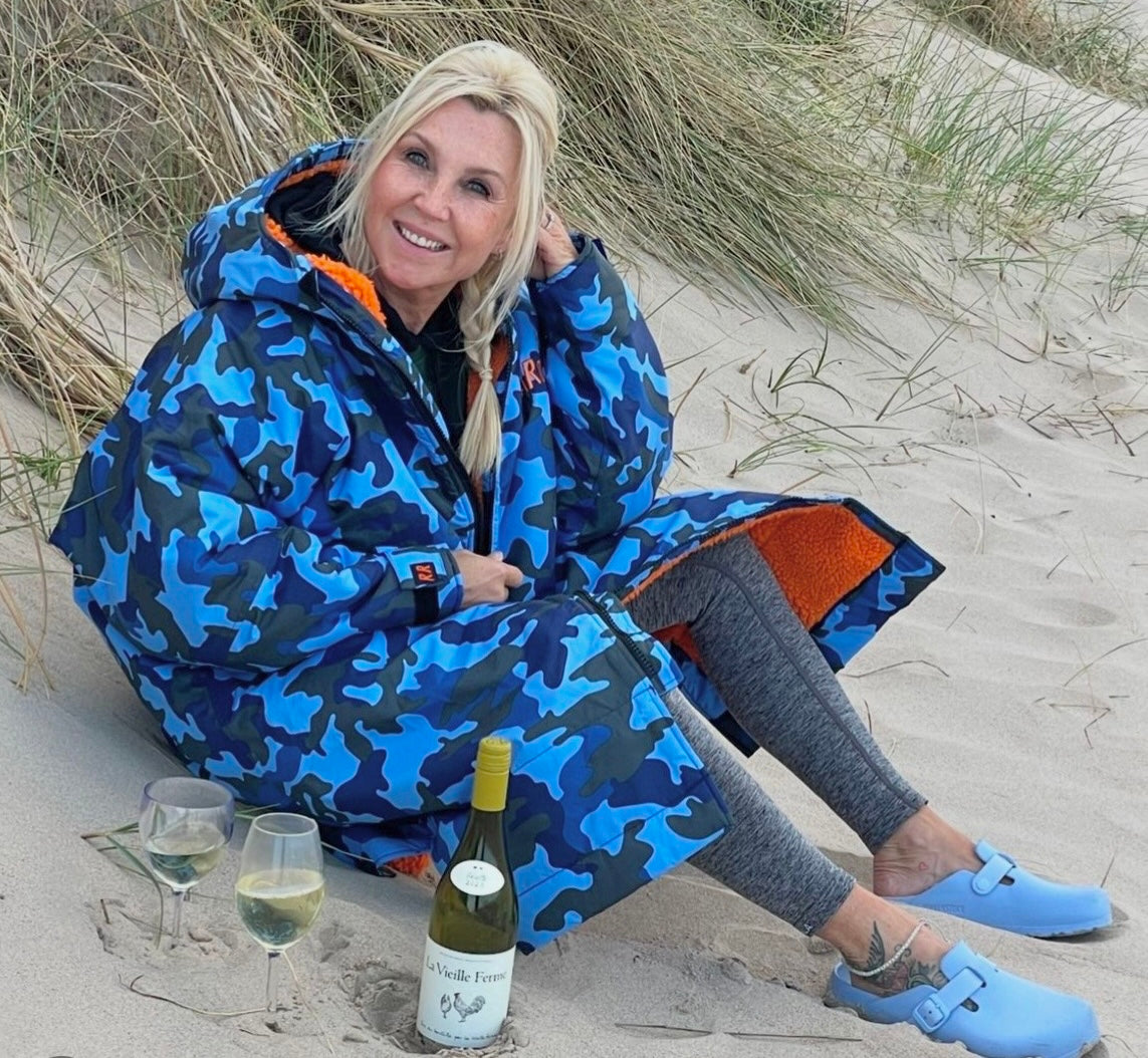 A woman in a blue camouflage robe is sitting on the sand with a bottle of wine and glasses.