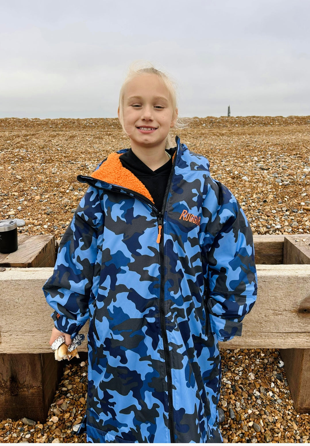 Child wearing a blue camouflage ruggarobe with orange lining on a beach.