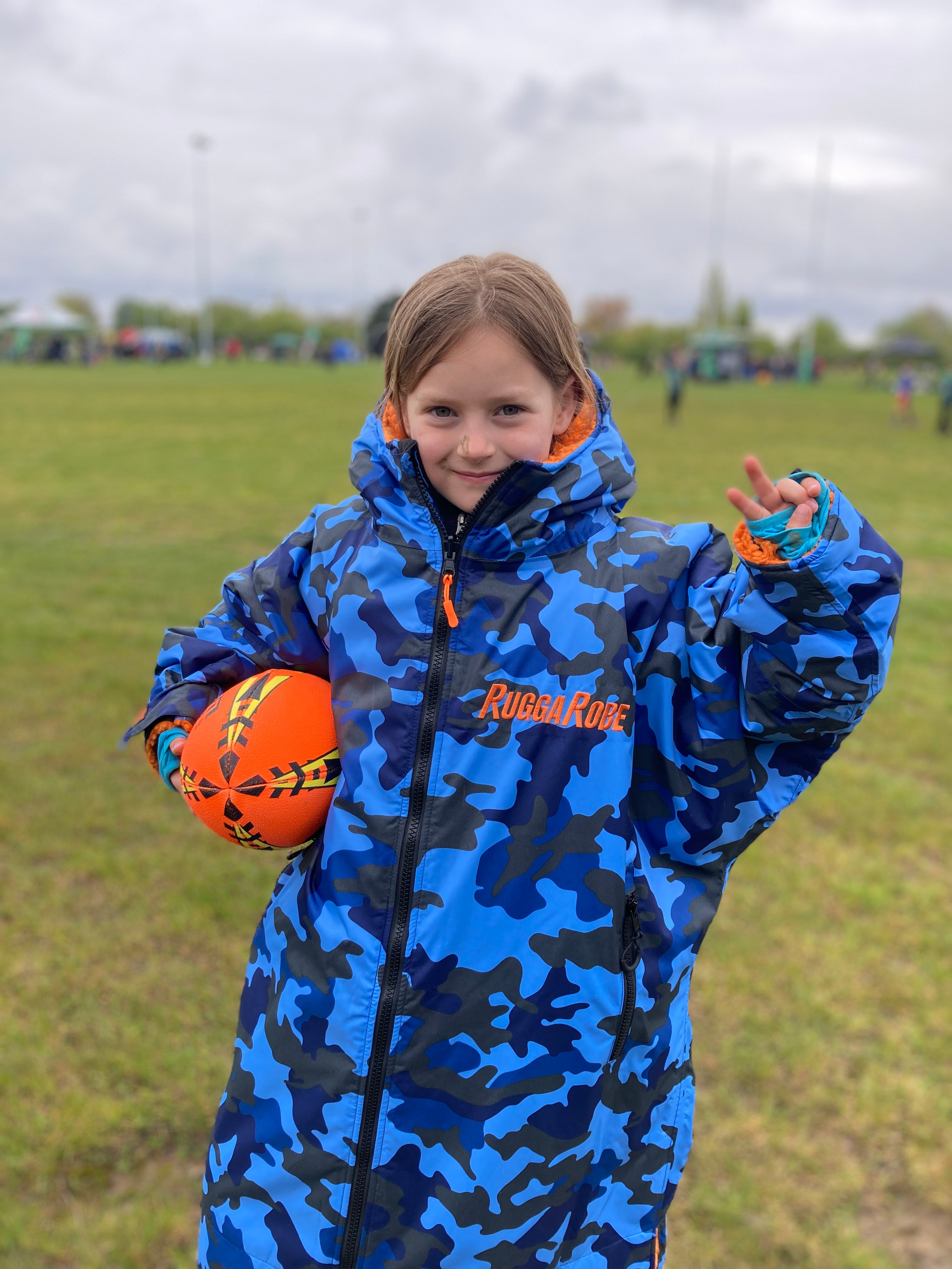 A child wearing a blue camouflage change robe with orange fleece lining, holding an orange ball, standing outdoors.