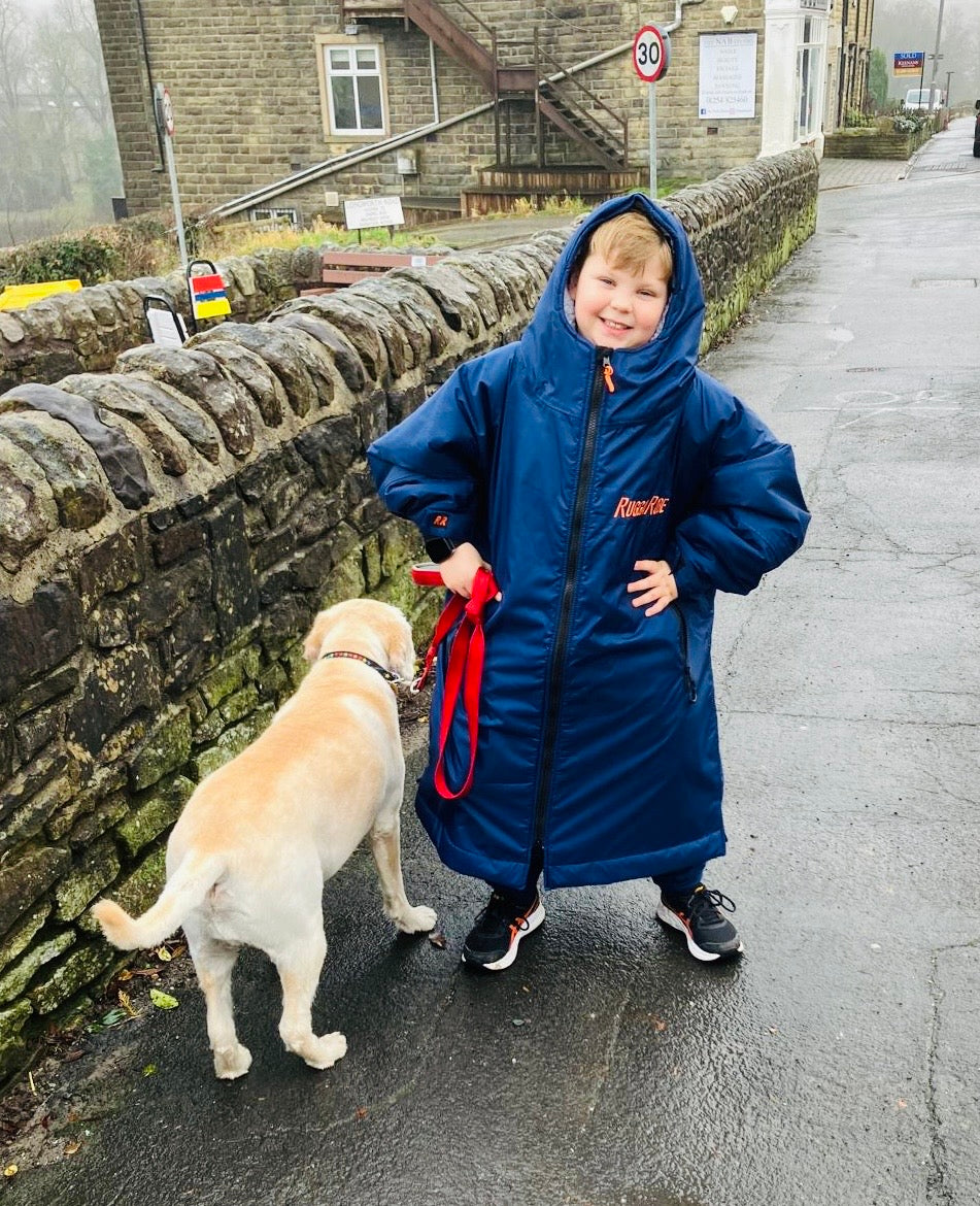 Child in a blue RuggaRobe with a dog on a leash near a stone wall.