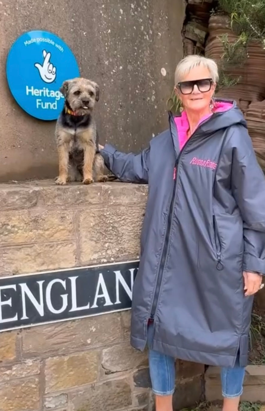 Woman in a long coat standing next to a dog with a 'Heritage Fund' sign in the background.