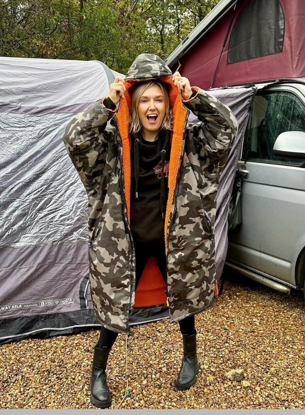 A person wearing a camouflage change robe with orange trim in front of a tent and a car.