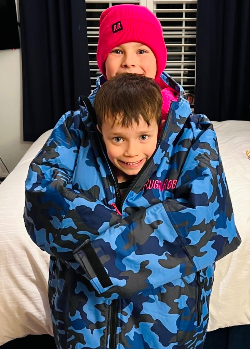Two children wearing blue camouflage ruggareobes with visible brand logos indoors.