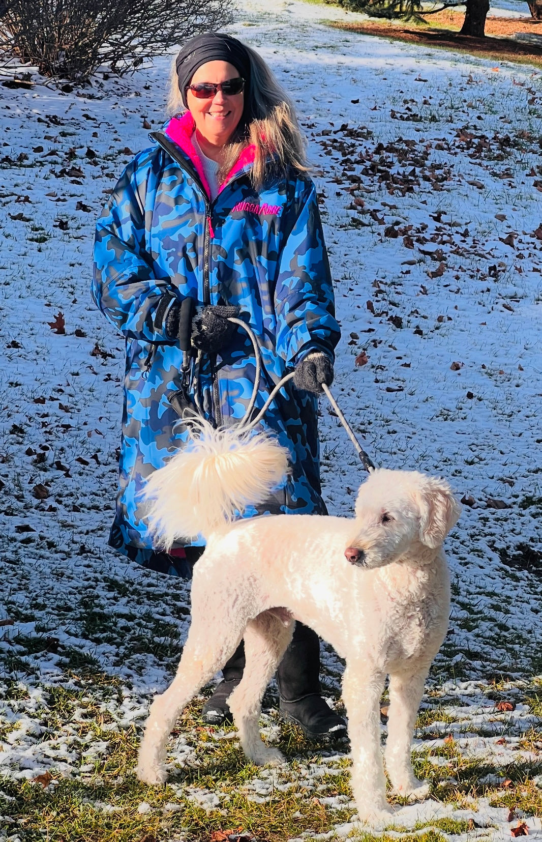 Woman in a blue camo ruggarobe walking a white dog on a snowy path