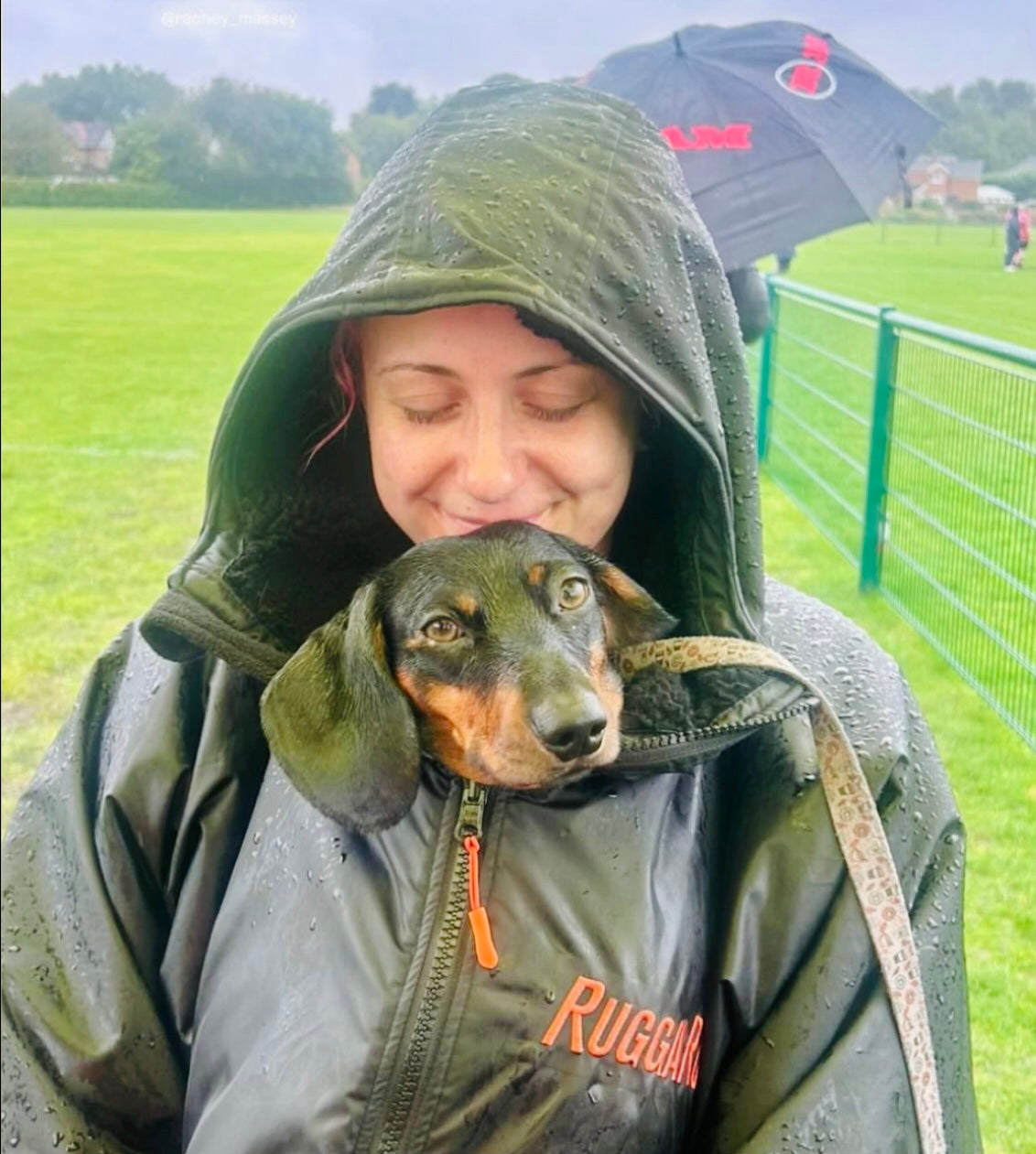Person in a ruggarobe with a dog peeking out from under the hood in a grassy field.