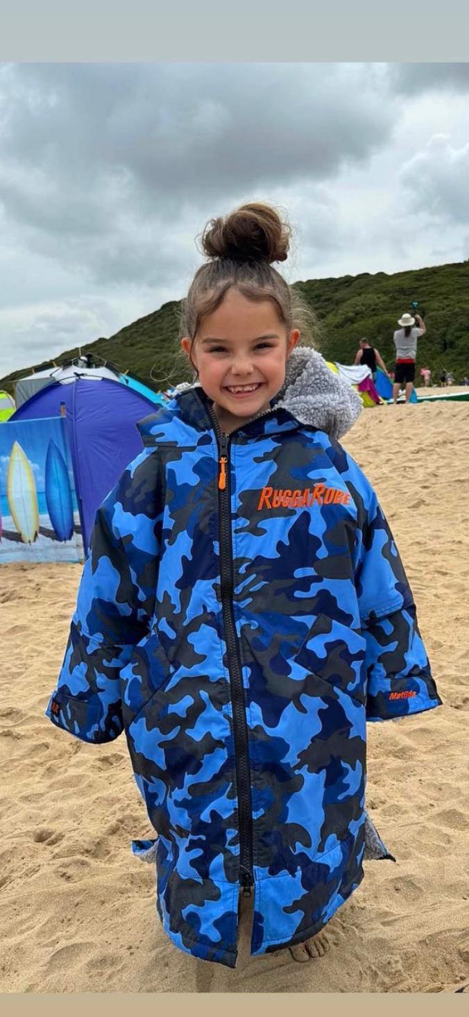 Child wearing a blue camouflage ruggarobe on a sandy beach with a tent and people in the background.