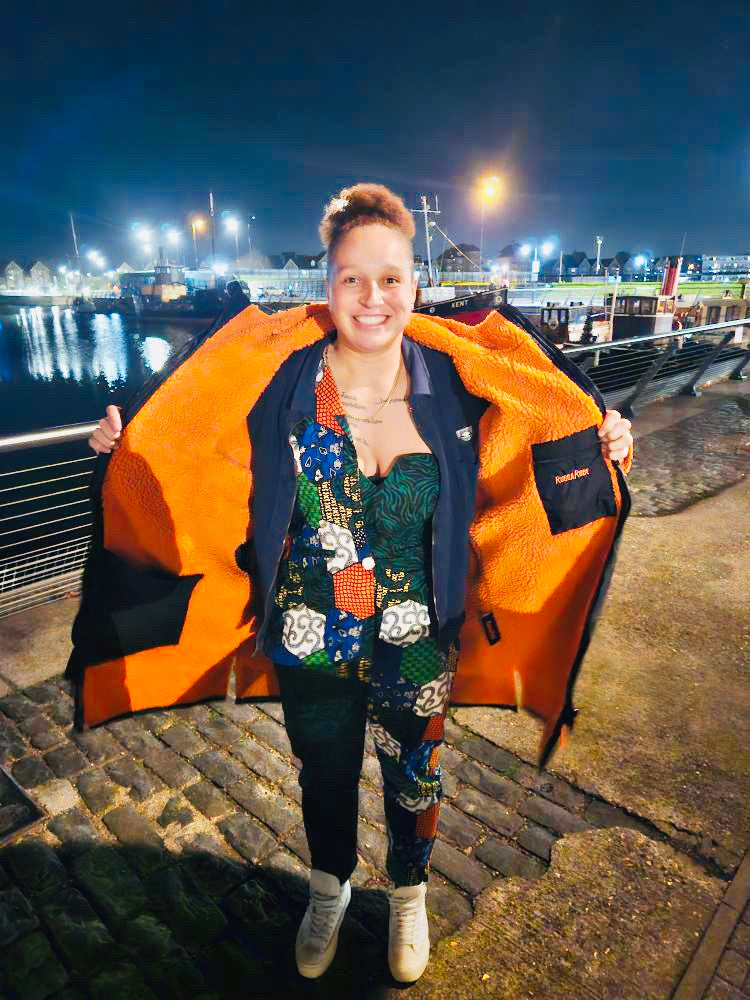 Woman wearing a black changerobe with orange lining and 'RuggaRobe' branding. Standing on a harbour