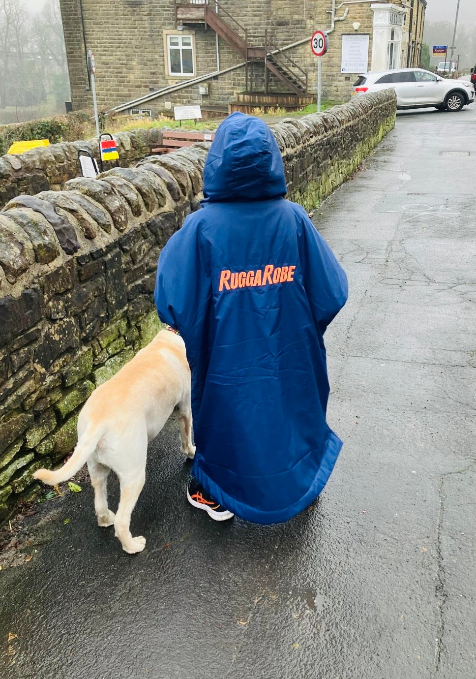 Child wearing a blue change robe with 'Ruggarobe' branding, walking a dog on a wet street.