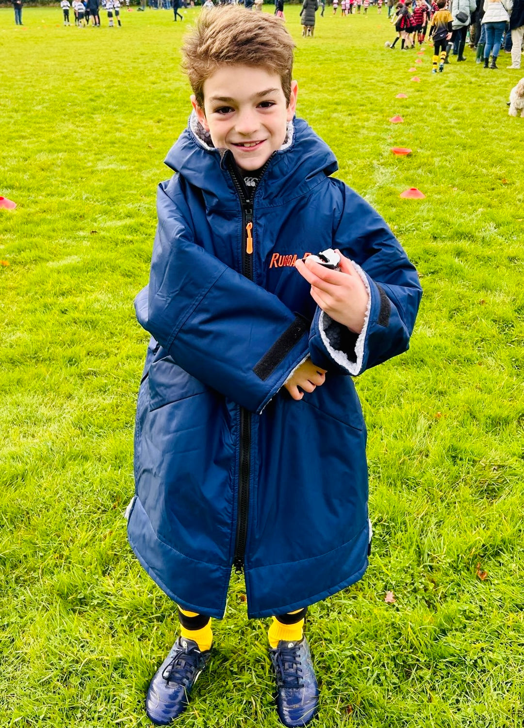 Child in a blue ruggarobe holding a device on a grassy field with people in the background