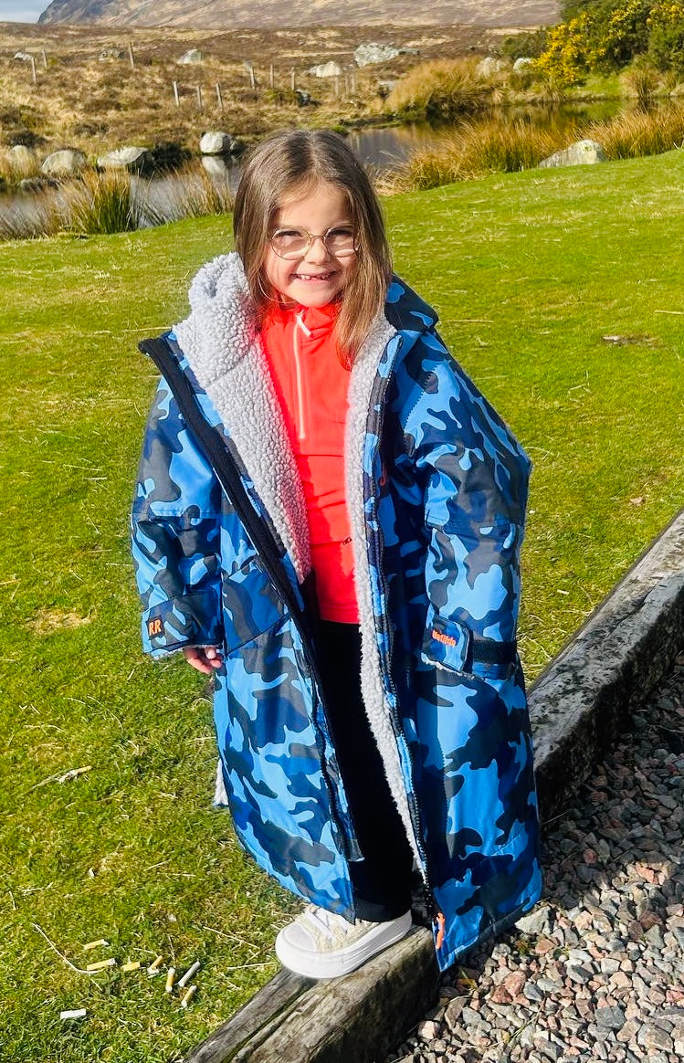 Child wearing a blue camouflage ruggarobe standing outdoors near a grassy area with a pond.
