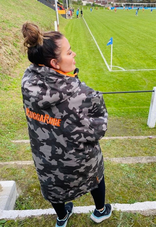 Person wrapped in a camouflage blanket with 'RuggaRobe' branding on a sports field.