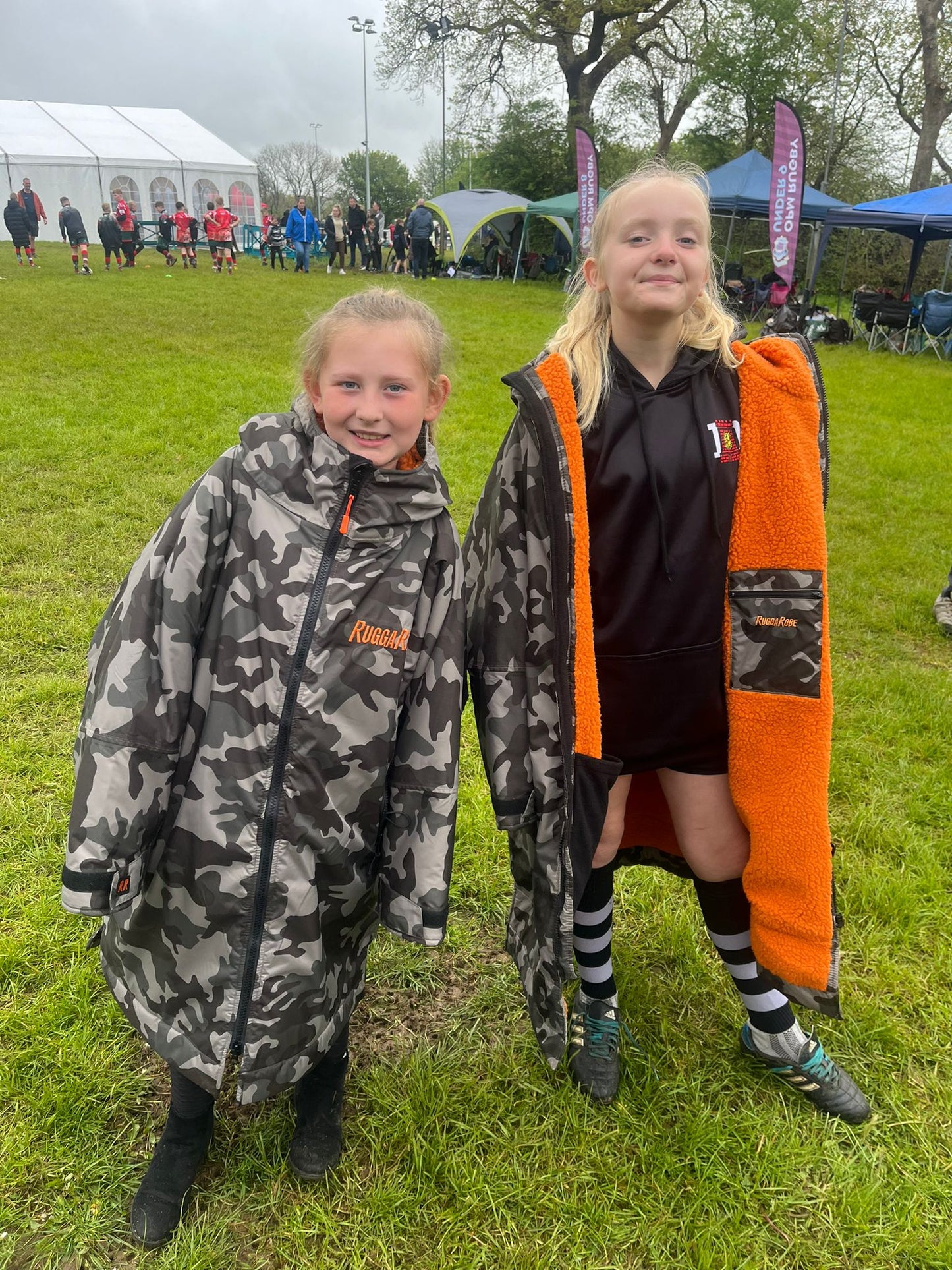 Two children wearing RuggaRobe change robes with camouflage patterns and orange accents on a sports field
