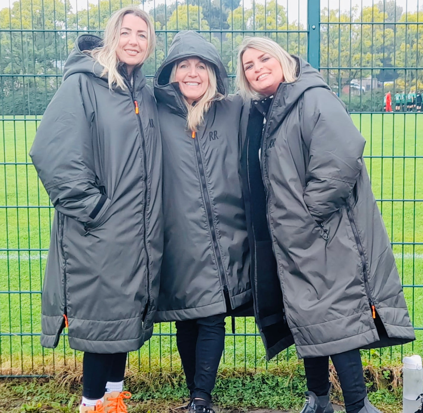 Three women wearing matching Green RuggaRobesoutdoors with a green fence at the football pitches 