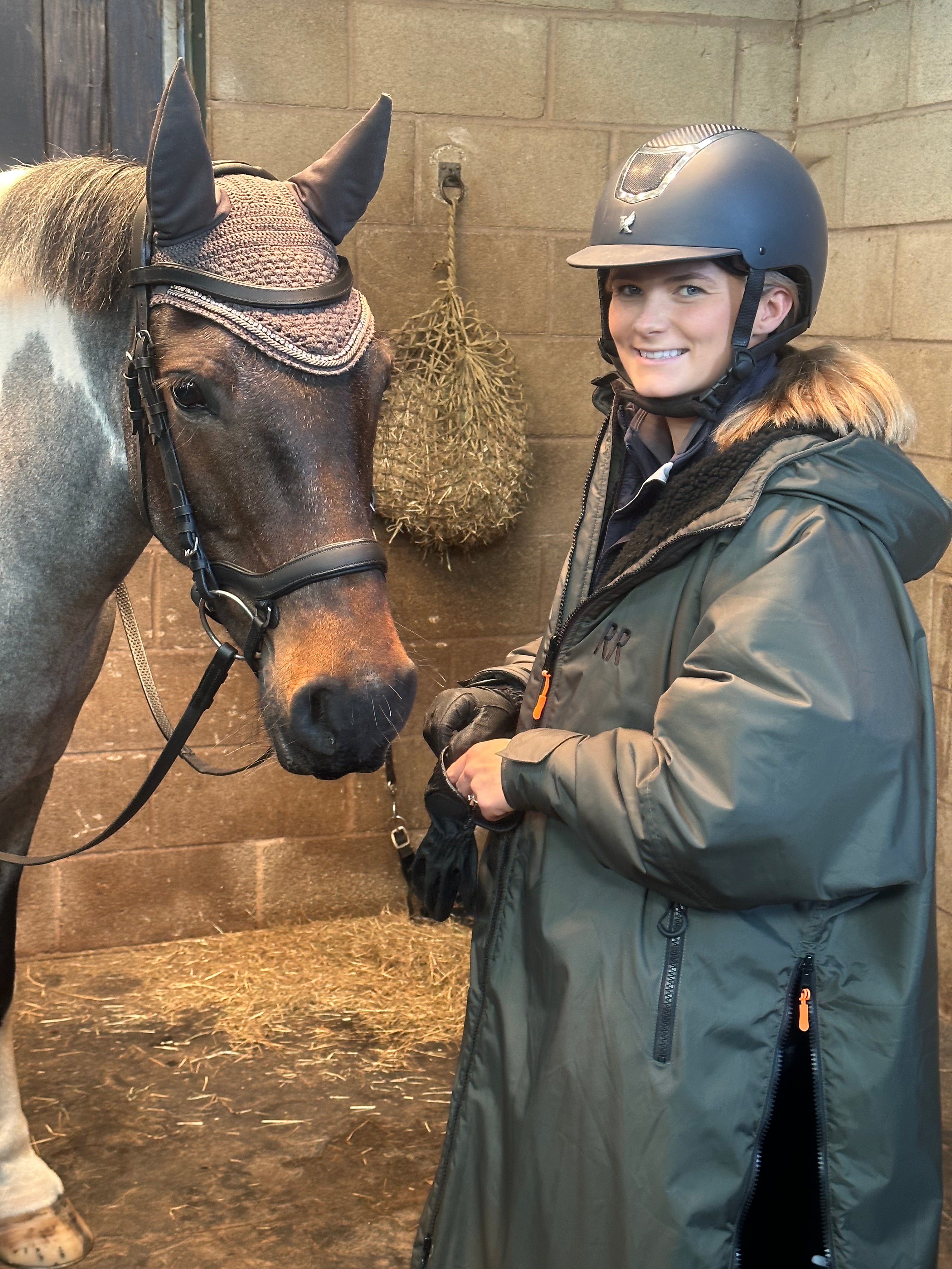A person standing next to a horse, wearing a green country robe with black fleece lining, featuring a hood and external pockets.