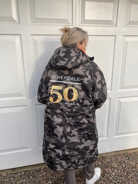 Person wearing a camouflage jacket with 'Emmerdale' branding in front of a white door.