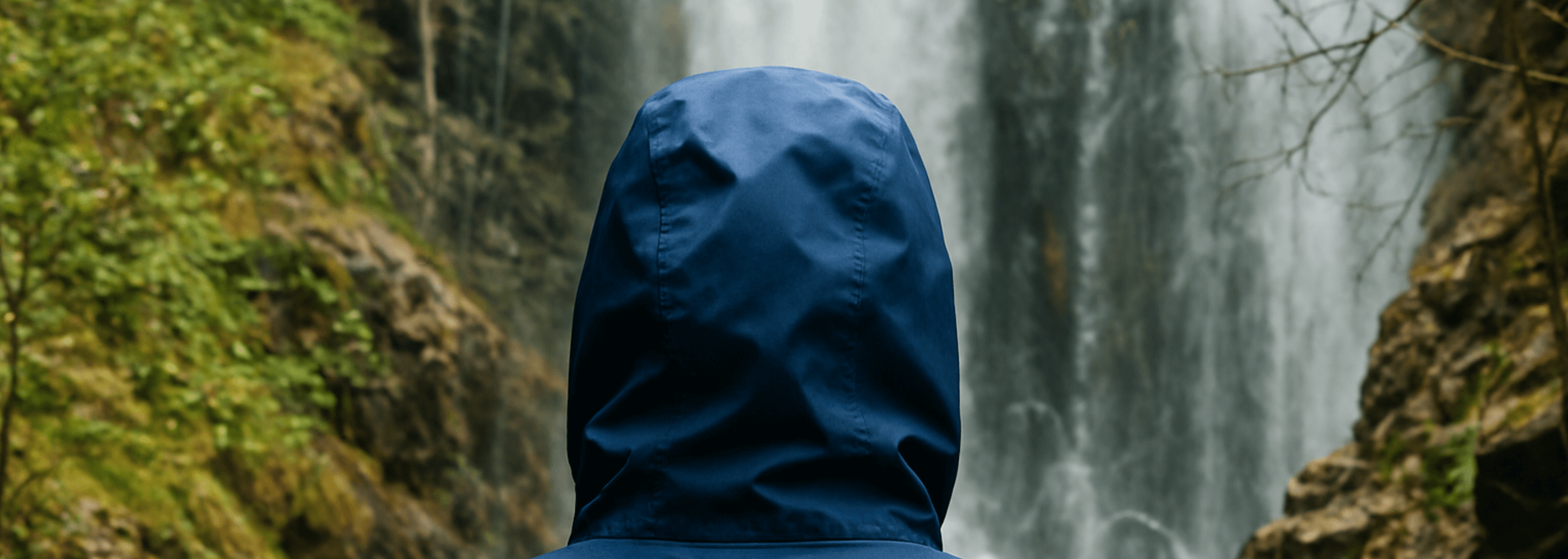 Person wearing a blue hooded jacket standing in front of a waterfall.